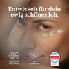 Eine lächelnde Person mit zwei Cremepunkten unter dem Auge. Der Text auf Deutsch lautet „Entwickelt für dein ewig schönes Ich“. Unten sind verschiedene Zertifizierungslabels abgebildet. Produktname: Hyaluron-Augencreme Markenname: Nature Love
